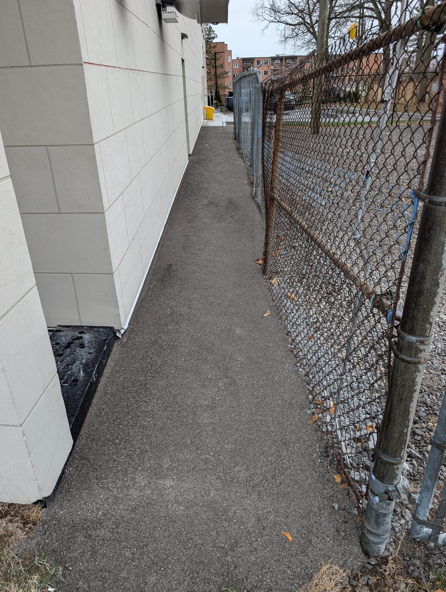 Narrow paved walkway between building and chain-link fence.