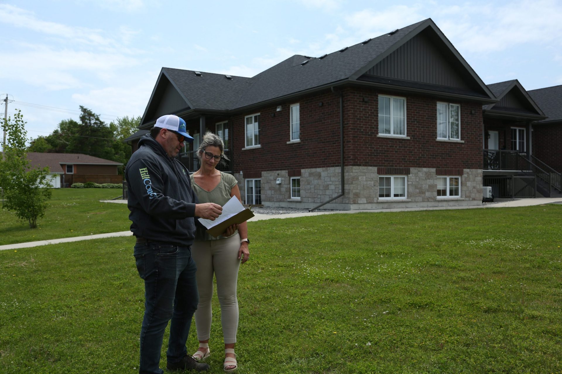 A man wearing a grey "Hill County" hoodie and a baseball cap stands on a green lawn next to a woman. He is holding a clipboard and pointing at a document, appearing to discuss details with her. In the background, there is a large, modern dark-brick two-story house with light stone accents along the foundation and several white-trimmed windows.